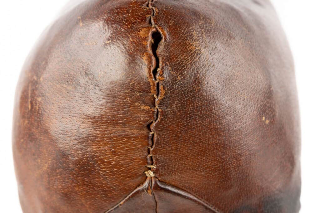 Close-up van een oude, versleten bruine leren voetbal met opvallende stiksels en zichtbare slijtplekken op het oppervlak.