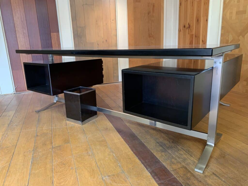 Een modern zwart bureau met metalen poten en twee grote open opbergvakken eronder, op een houten vloer met diverse houten panelen op de achtergrond.