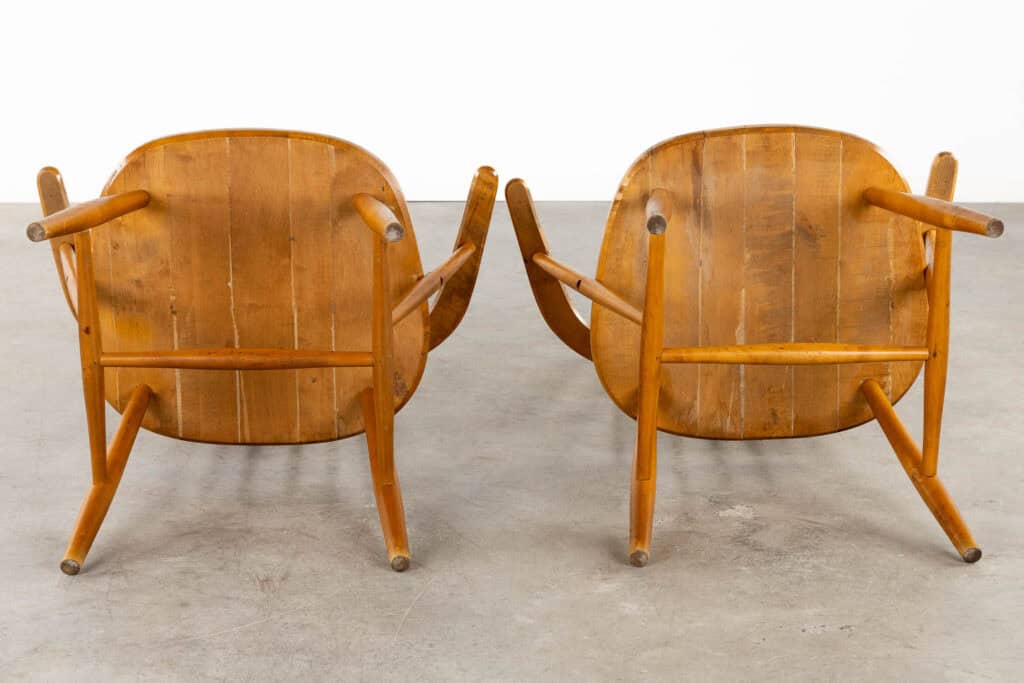 Twee houten stoelen met gebogen rugleuningen en armleuningen staan rug aan rug op een betonnen vloer, van achteren gezien. De stoelen hebben een mid-century modern design met zichtbare houtnerf.