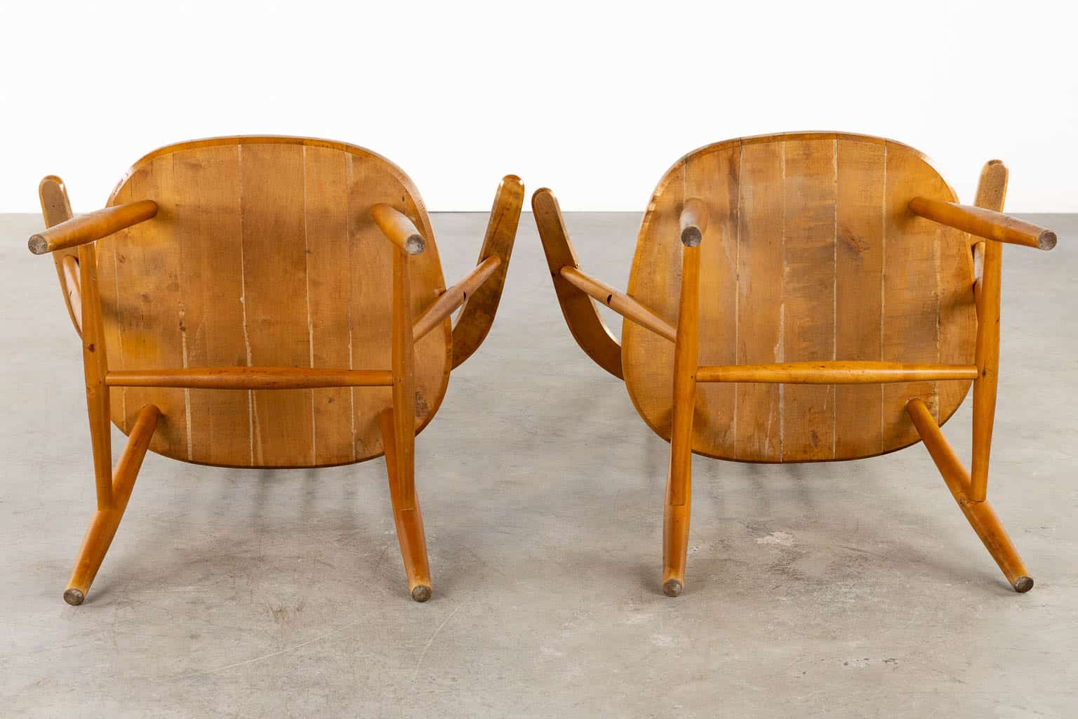 Twee houten stoelen met gebogen rugleuningen en armleuningen staan rug aan rug op een betonnen vloer, van achteren gezien. De stoelen hebben een mid-century modern design met zichtbare houtnerf.