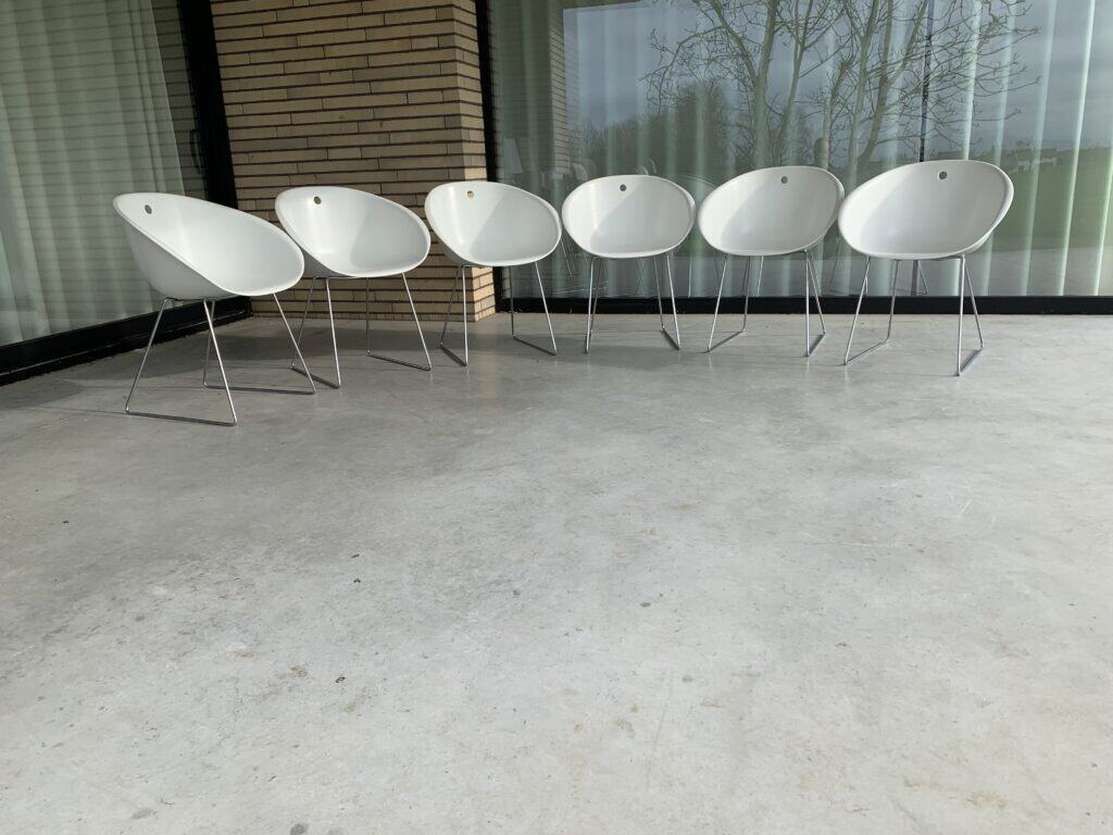 Zes moderne witte stoelen met metalen poten staan op een rij op een betonnen terras voor een glazen wand en een hoek van baksteen, waarin de bomen en een buitentafereel worden weerspiegeld.