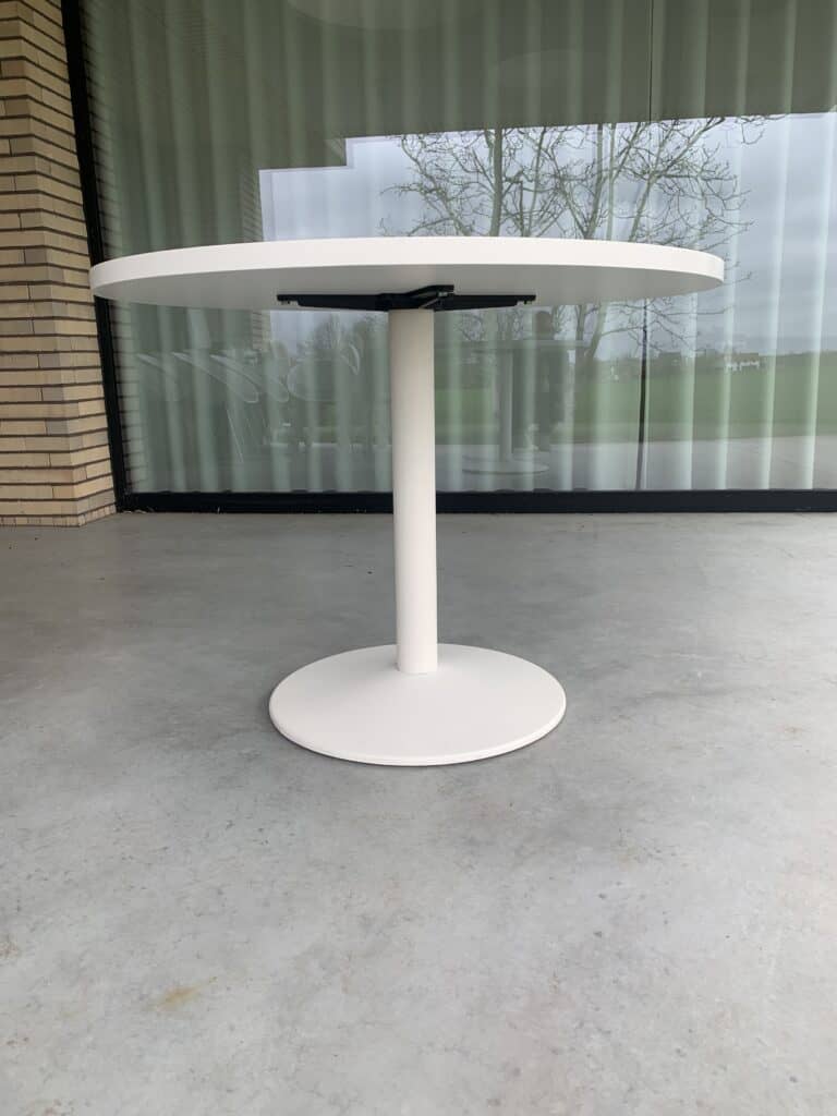 Een moderne, ronde, witte tafel met een enkele centrale voet staat op een gladde, grijze betonnen vloer, met een groot glazen raam en een bakstenen muur op de achtergrond.