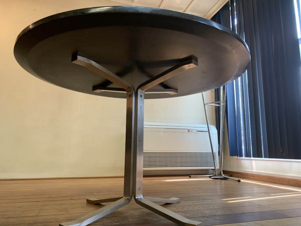 Een ronde zwarte tafel met een metalen voetstuk staat op een houten vloer bij een raam met verticale jaloezieën. Op de achtergrond zijn een airconditioning en een hoge metalen standaard zichtbaar.