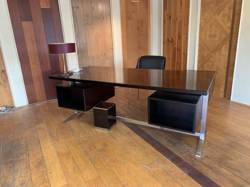 Een modern bureau met een donkerhouten oppervlak, open planken, een bijpassende prullenbak, een kastanjebruine bureaulamp en een zwarte stoel, op een houten vloer in een kamer met houten lambrisering.