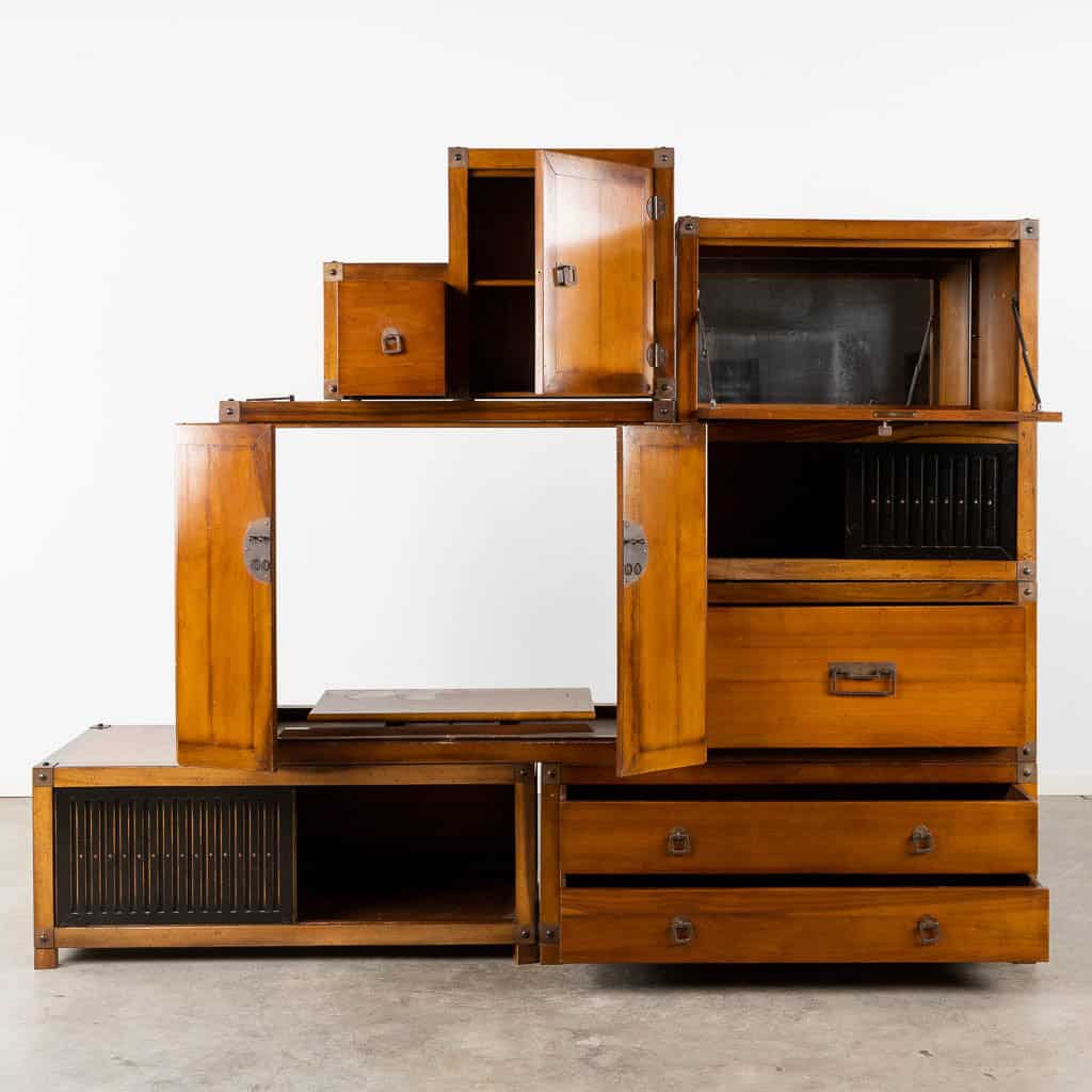 Een modulaire houten kast in vintagestijl met verschillende compartimenten, lades en deuren, gerangschikt in een asymmetrische stapel tegen een witte muur op een betonnen vloer.
