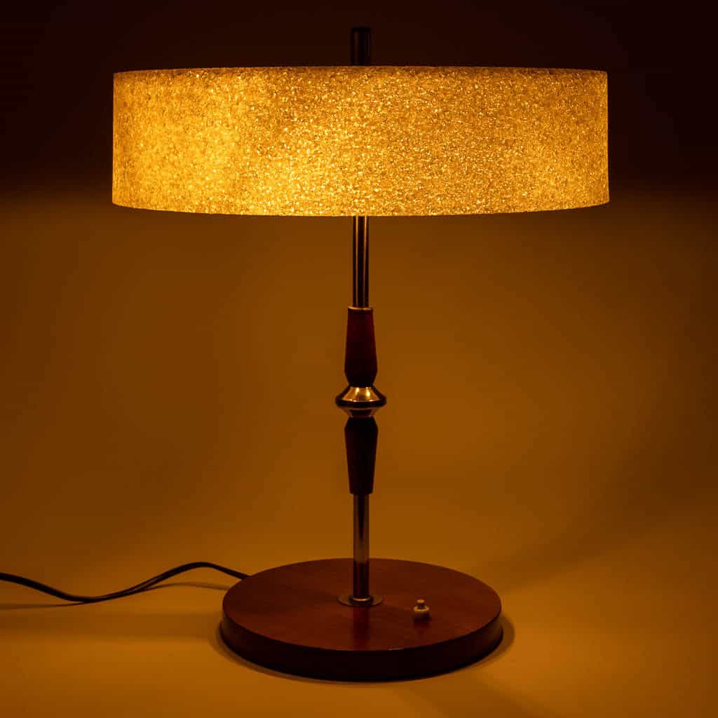 Een vintage tafellamp met een ronde, getextureerde gele lampenkap straalt een warme gloed uit. De lamp heeft een houten en metalen steel, een ronde houten voet en een zichtbaar zwart snoer op een effen achtergrond.
