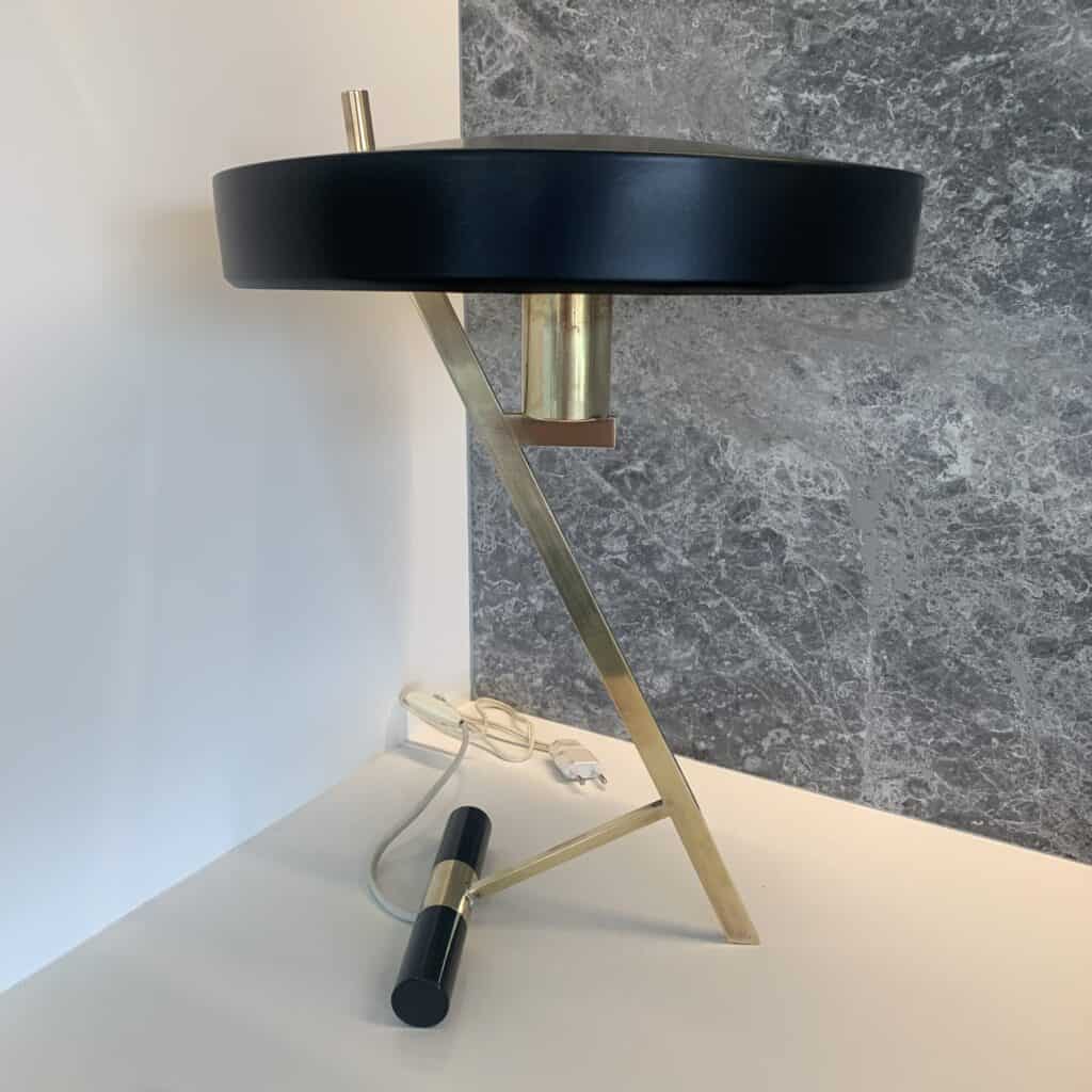 Een moderne tafellamp met een zwarte ronde kap en een goudkleurige metalen voet, met een minimalistisch geometrisch ontwerp. De lamp staat op een witte ondergrond tegen een grijze stenen muur, met het snoer en de stekker zichtbaar.