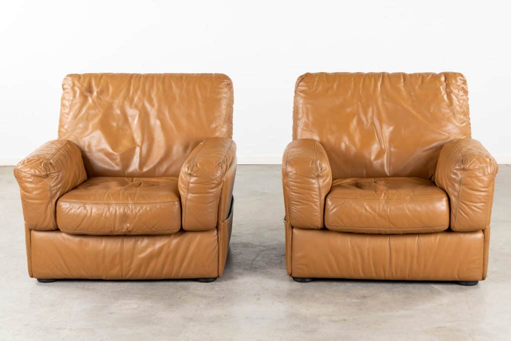 Twee bruine leren fauteuils met brede zitkussens en licht gekreukte bekleding staan naast elkaar op een betonnen vloer tegen een eenvoudige witte muur.