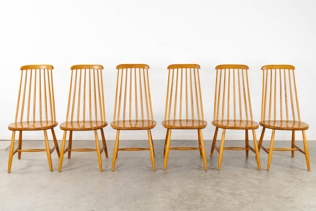 Zes houten stoelen met spijlenleuningen staan netjes op een rij tegen een eenvoudige witte muur op een lichte betonnen vloer.