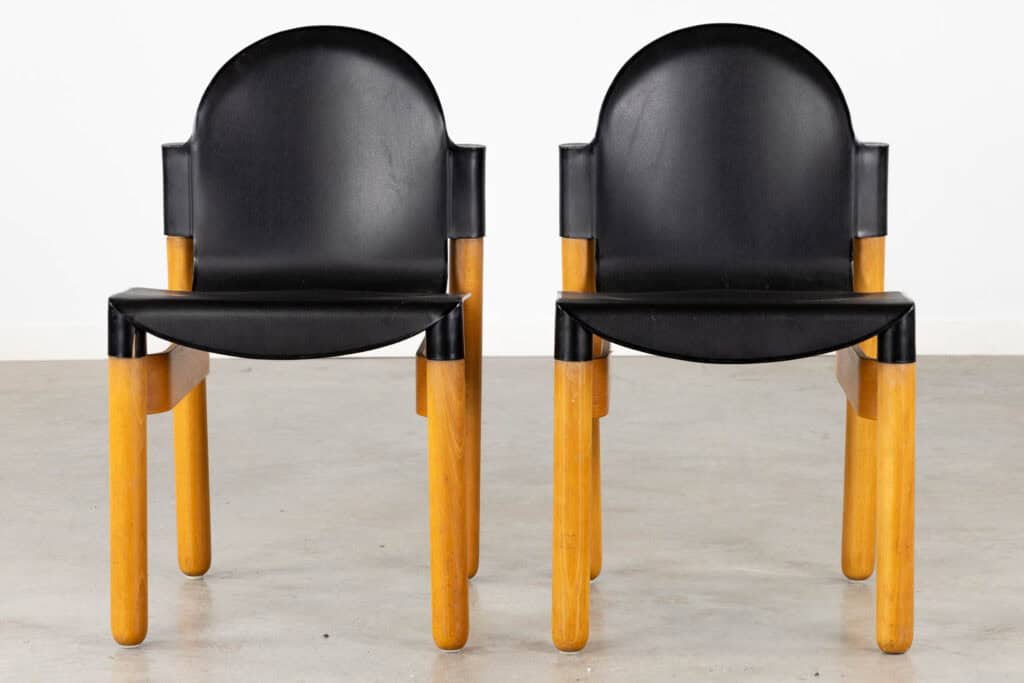 Twee moderne stoelen met zwarte, gebogen rugleuningen en zittingen en dikke, rechte houten poten staan naast elkaar op een gladde, grijze vloer tegen een eenvoudige witte muur.