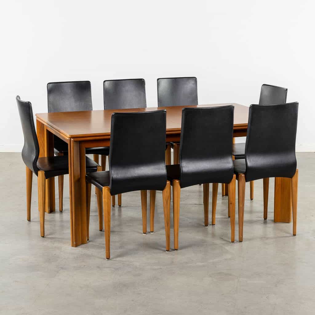 Een rechthoekige houten eettafel, omringd door acht zwarte stoelen met hoge rugleuningen en houten poten, op een lichtgrijze vloer tegen een effen witte muur.