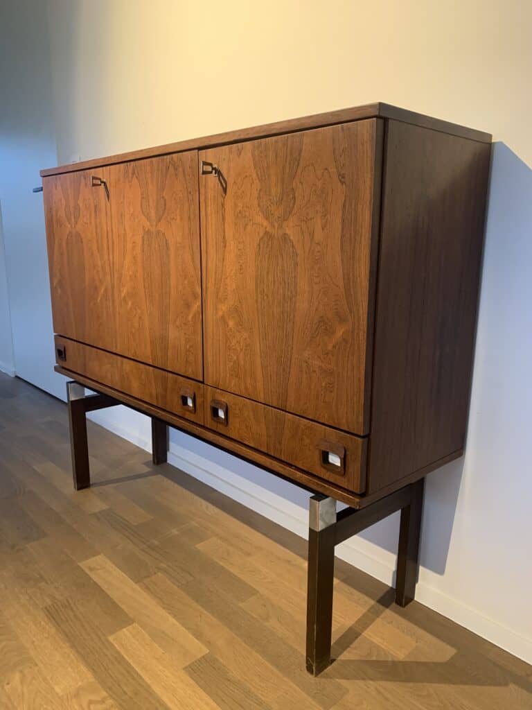 Een houten kast in mid-century modern-stijl met twee deuren en twee kleine lades, met opvallende houtnerfpatronen, op hoge, donkere poten tegen een lichtgekleurde muur en houten vloer.