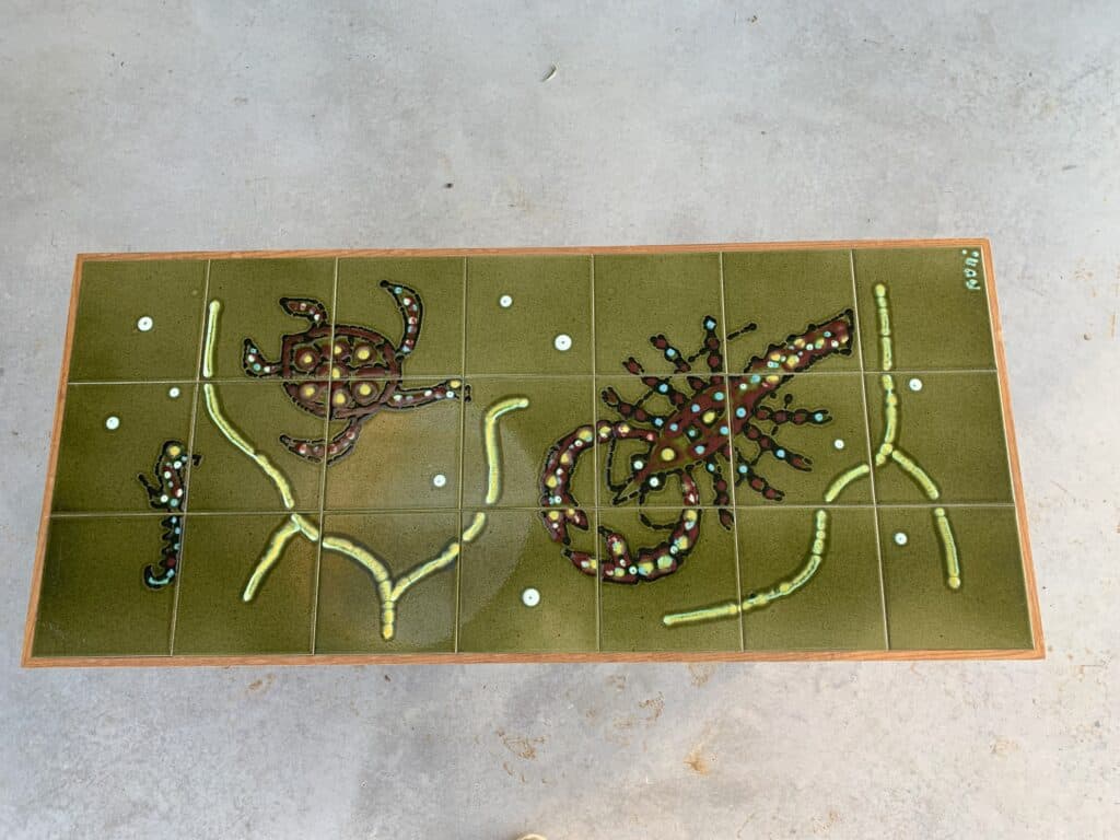 Een rechthoekig kunstwerk met punt- en lijnpatronen in Aboriginal-stijl, met een schildpad aan de linkerkant en een hagedis aan de rechterkant, tegen een olijfgroene achtergrond, gescheiden door tegellijnen.