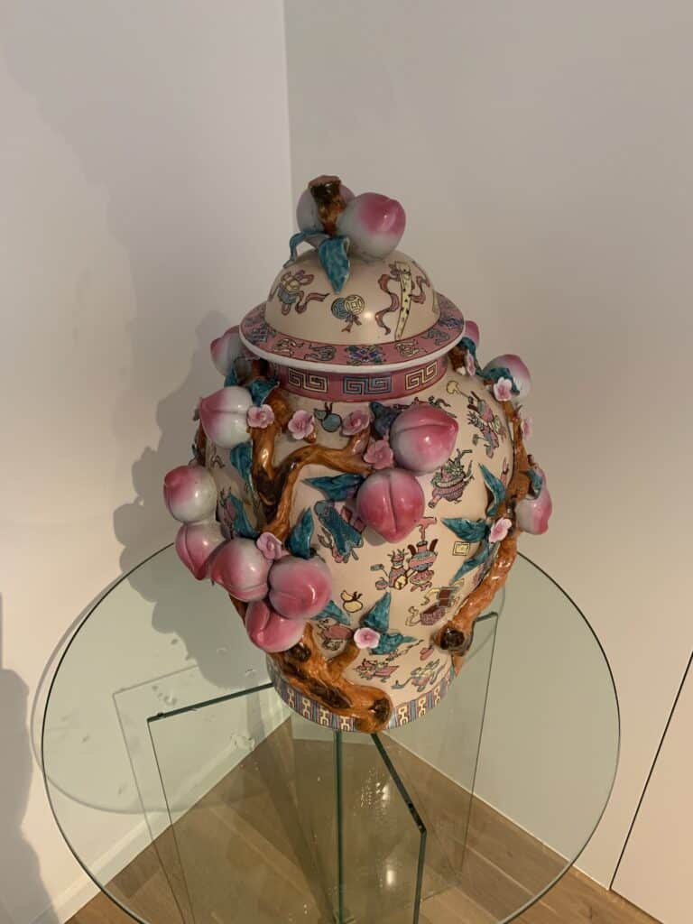 Een decoratieve keramische vaas met deksel, versierd met roze perzikornamenten, takken en geschilderde figuren, staat op een tafel met glazen blad in de hoek van een kamer met lichte muren en een houten vloer.