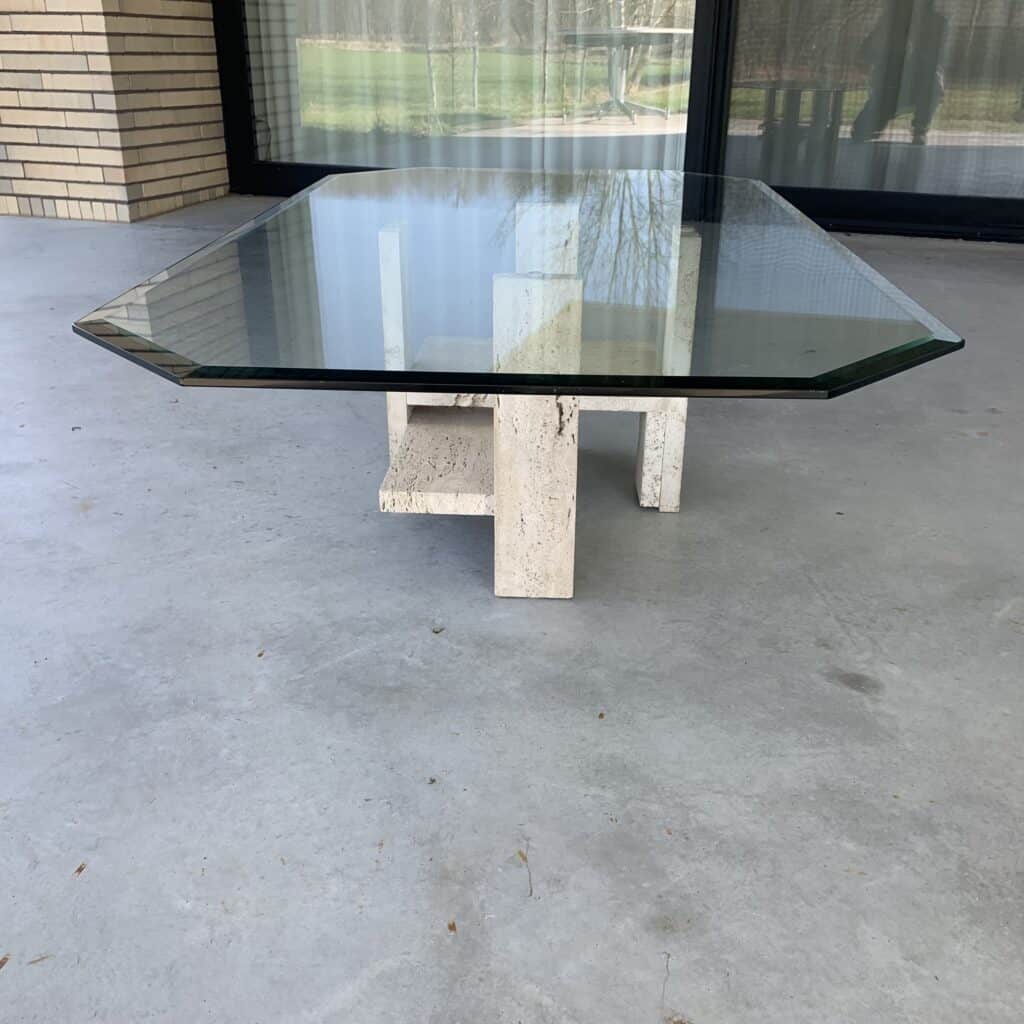 Een moderne salontafel met een dik, rechthoekig glazen blad en een geometrisch stenen onderstel staat op een grijze betonnen vloer, vlak bij een groot raam en een bakstenen muur. Buiten zijn bomen en gras zichtbaar.