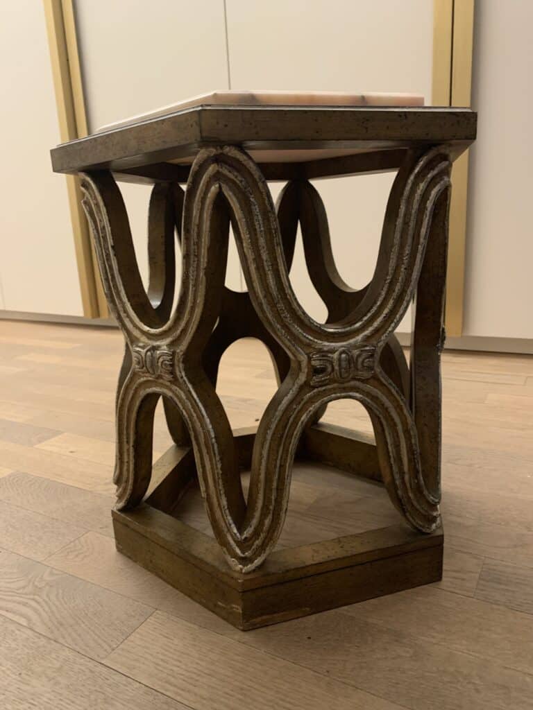 Een zeshoekige houten kruk met een ingewikkeld, golvend, in elkaar grijpend ontwerp aan de zijkanten. De kruk heeft een metallic bronzen afwerking met witte accenten en staat op een lichte houten vloer.