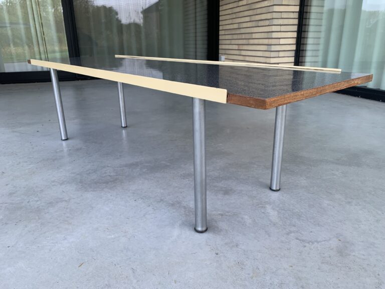 Een moderne rechthoekige tafel met een donker oppervlak, een houten rand en vier metalen poten staat op een grijze betonnen vloer voor grote ramen en een bakstenen muur. Twee lichtgekleurde houten latten rusten erop.