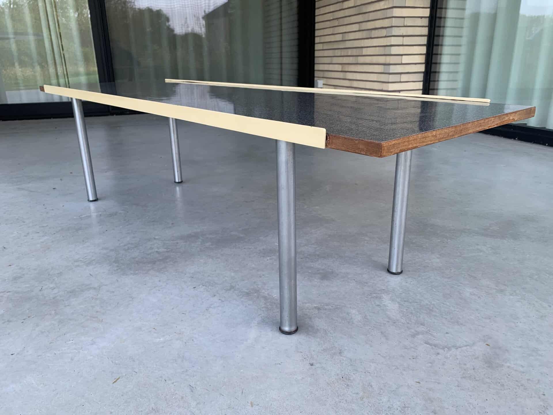 Een moderne rechthoekige tafel met een donker oppervlak, een houten rand en vier metalen poten staat op een grijze betonnen vloer voor grote ramen en een bakstenen muur. Twee lichtgekleurde houten latten rusten erop.