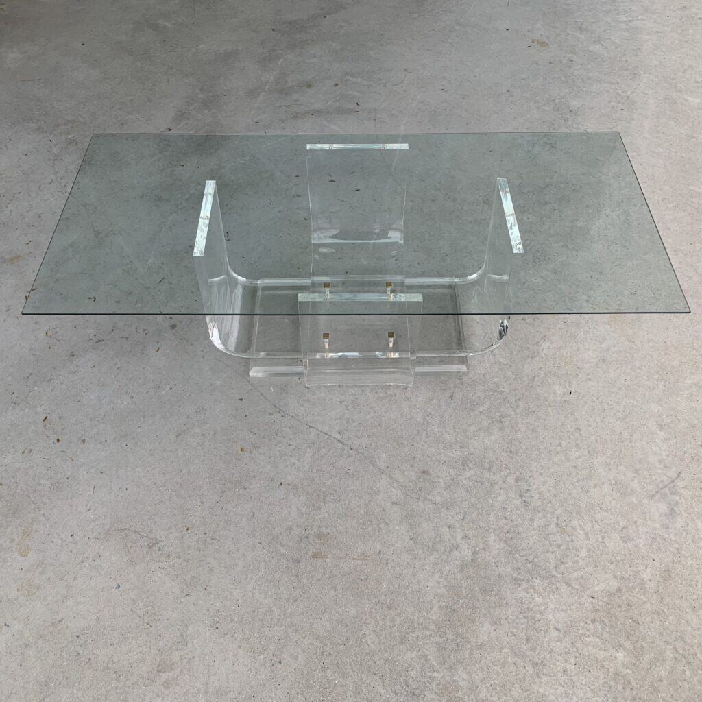 Een rechthoekige glazen salontafel met een transparant acryl onderstel, geplaatst op een lichte betonnen vloer. Het ontwerp is modern en minimalistisch, met strakke lijnen en heldere materialen.