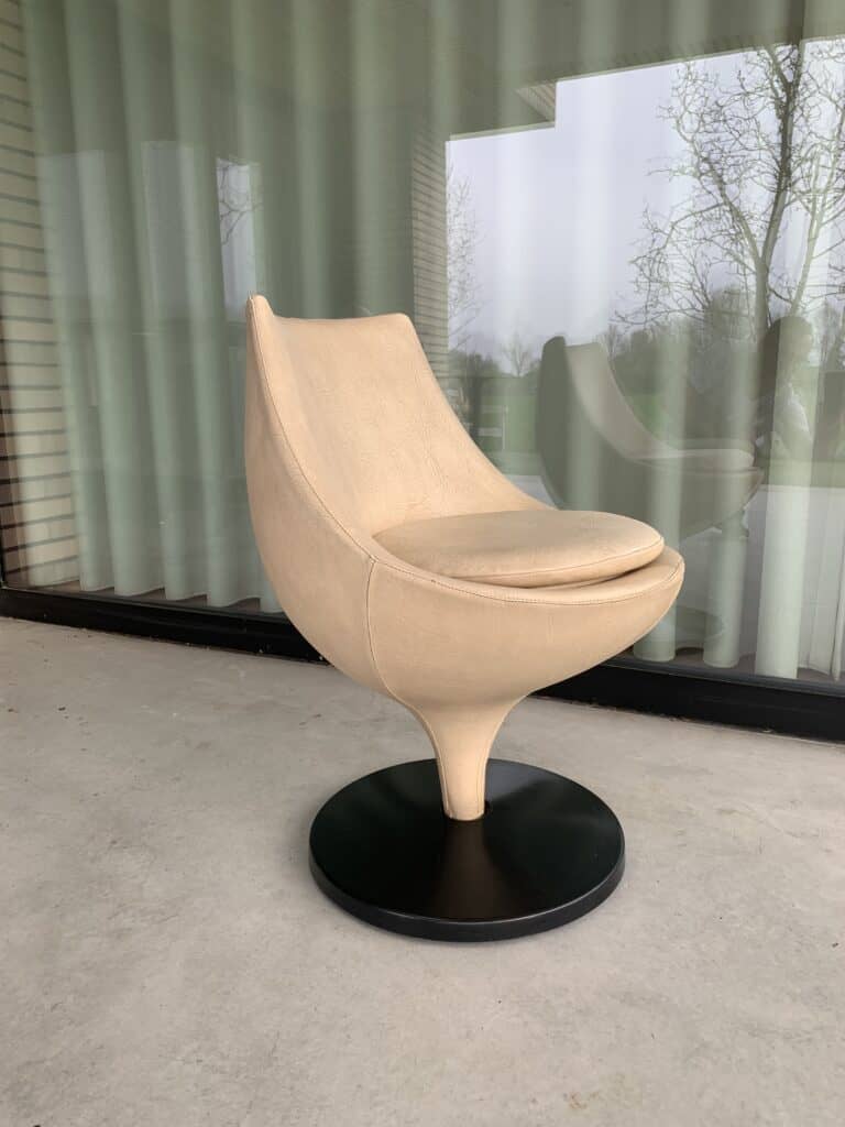 Een moderne beige draaistoel met een gebogen rugleuning en zitting op een zwarte ronde voet, geplaatst op een betonnen terras voor grote glazen deuren met transparante gordijnen. Buiten zijn bomen en weerspiegelingen zichtbaar.