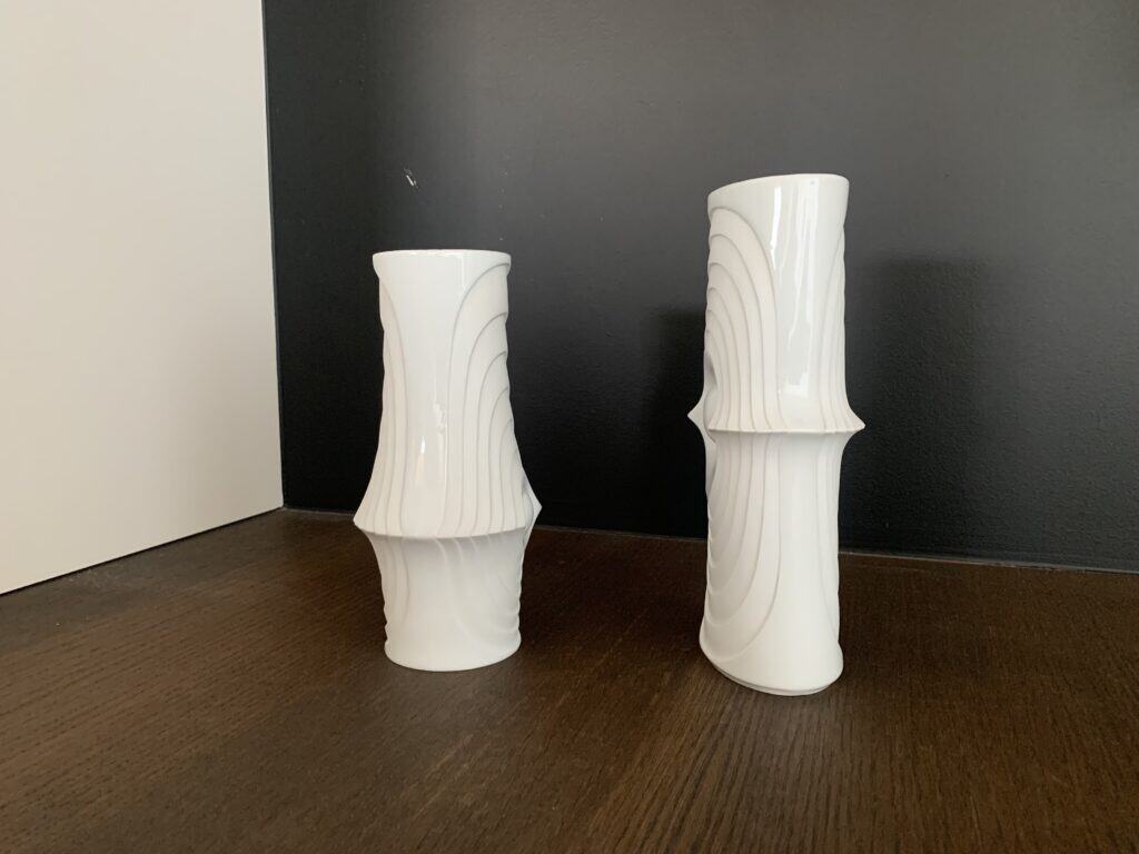 Twee witte keramische vazen met abstracte, golvende patronen staan op een donker houten oppervlak tegen een tweekleurige zwart-witte muur. De vazen hebben unieke vormen en een glanzende afwerking.