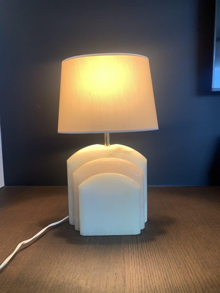 Een verlichte tafellamp met een beige kap en een witte voet in art-decostijl staat op een donker houten oppervlak tegen een donkere muur. Een wit snoer loopt uit de voet.
