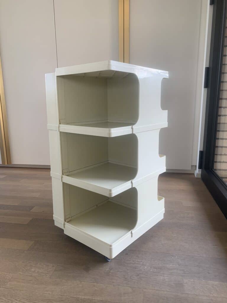 Een witte, drielaagse plastic opbergwagen met open zijkanten en afgeronde hoeken staat op een houten vloer bij een raam. Elke plank is ruim en de wagen staat op wielen voor mobiliteit.