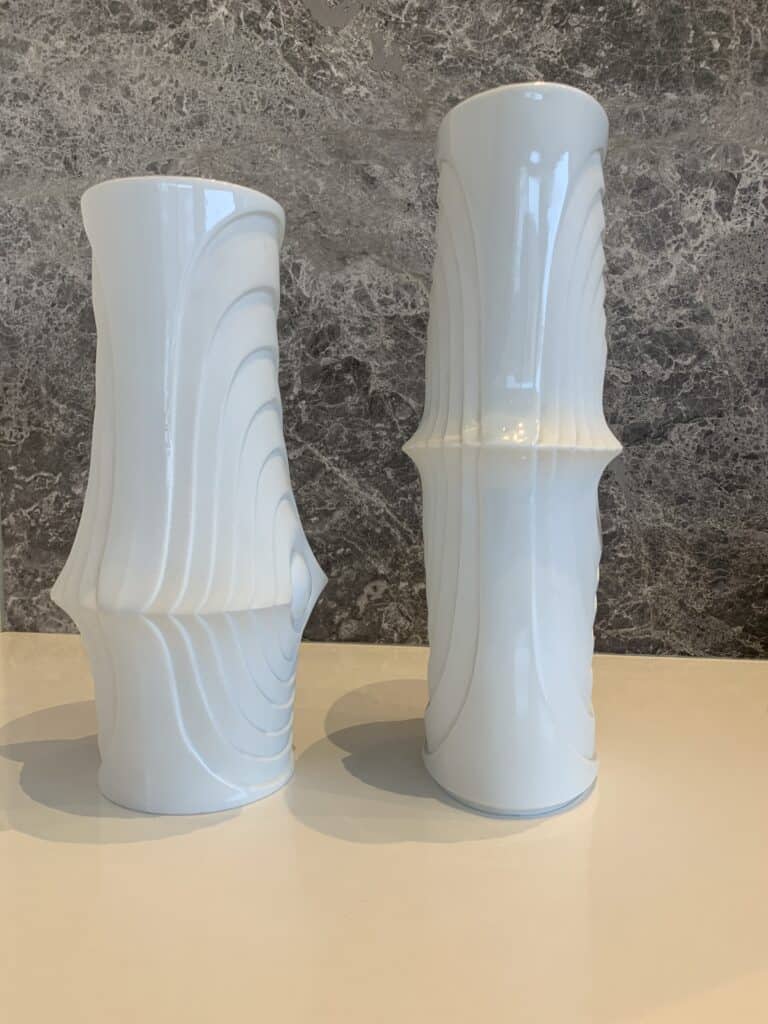 Twee hoge, glanzend witte keramische vazen met geometrische, gebogen randen staan naast elkaar op een wit oppervlak, tegen een donkergrijze achtergrond met marmerpatroon.
