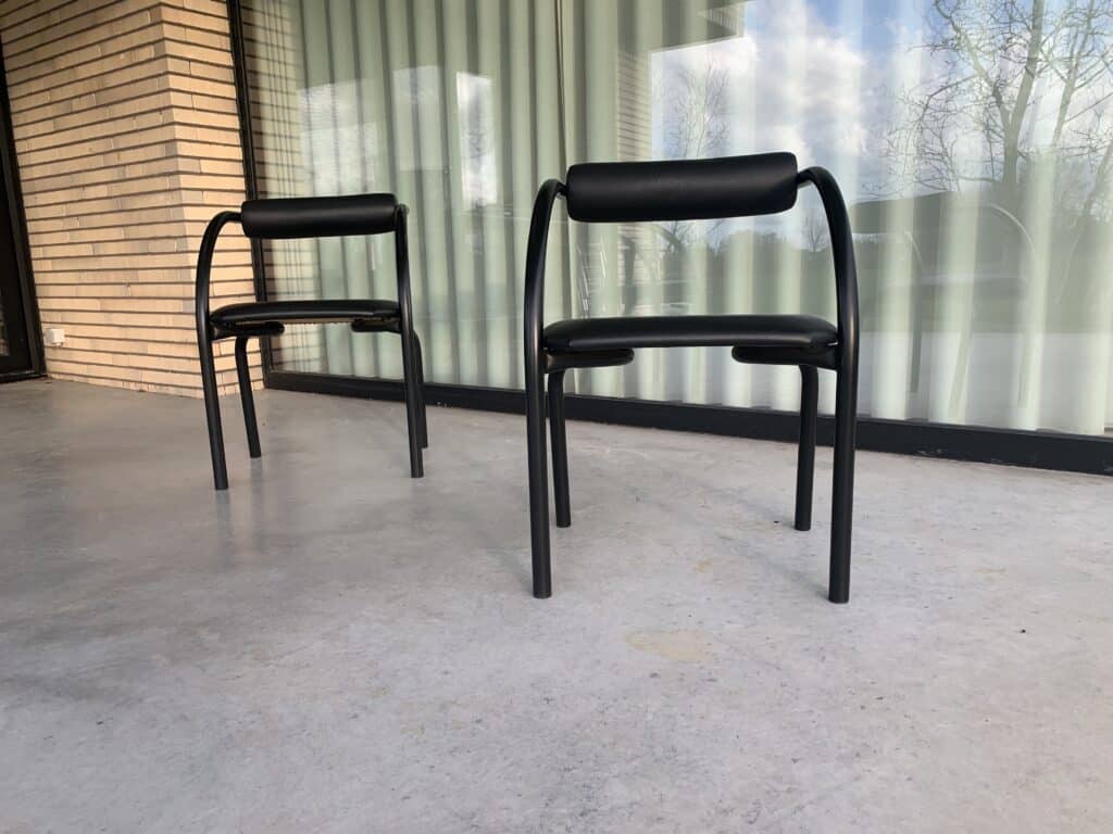 Twee zwarte stoelen met gebogen frame staan op een betonnen terras voor een glazen raam met verticale jaloezieën. Links is een bakstenen muur zichtbaar en de bomen weerspiegelen in het glas.