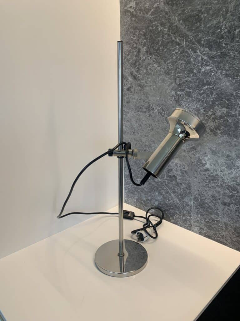 Een moderne, metalen, verstelbare bureaulamp met een cilindrische kop staat op een wit oppervlak bij een grijs gemarmerde muur. Het zwarte snoer ligt losjes opgerold op het bureau.