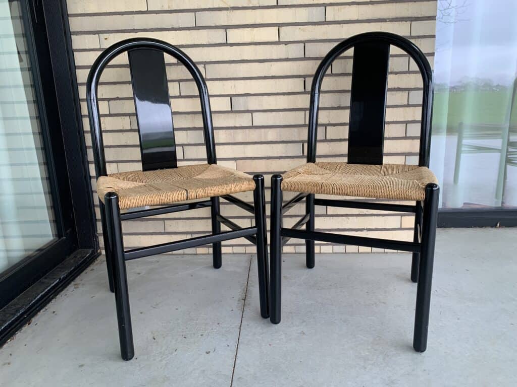 Twee zwarte metalen stoelen met gebogen rugleuningen en gevlochten rieten zittingen staan naast elkaar op een grijs terras, voor een lichte bakstenen muur en vlak bij grote glazen ramen.