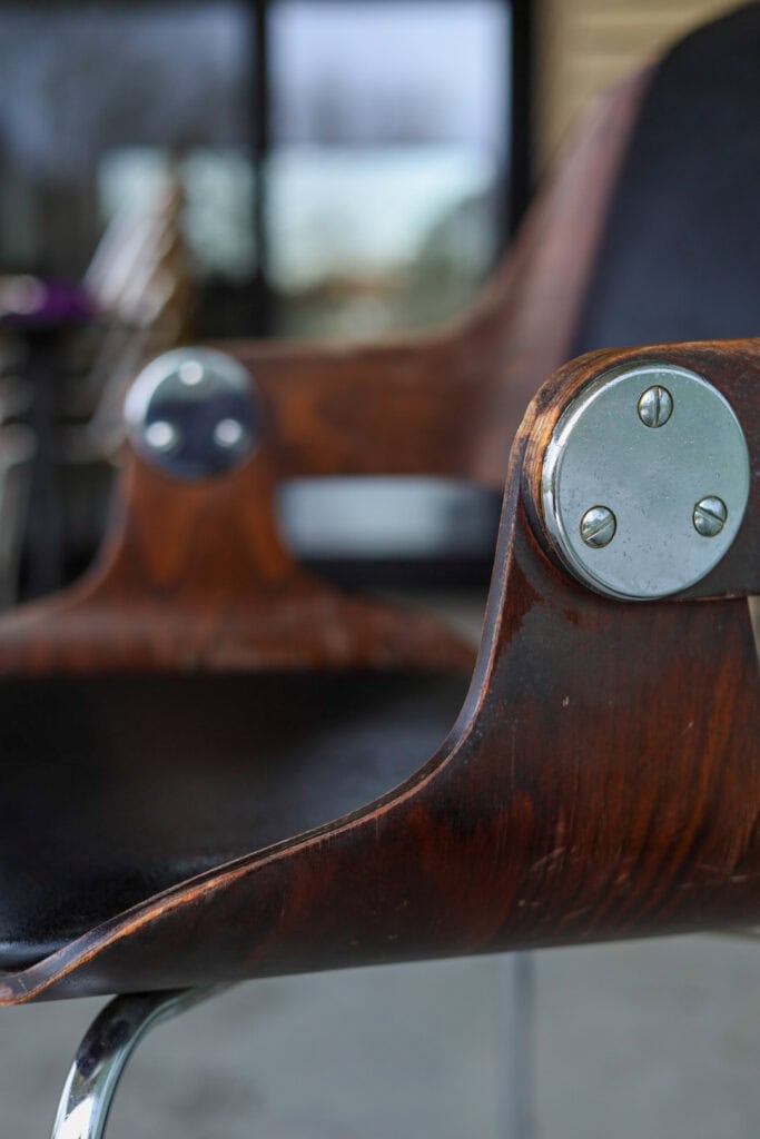 Close-up van een moderne houten stoel met armleuningen en metalen bevestigingsmiddelen, met een donkere afwerking en zwarte zitting, tegen een onscherpe achtergrond van ramen en buitenlandschap.