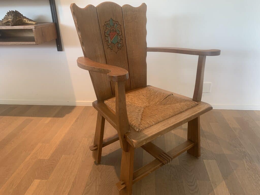 Een houten fauteuil met een gesneden en geschilderd bloemenmotief op de rugleuning en een gevlochten zitting, geplaatst op een lichte houten vloer bij een witte muur en een plank met een decoratief object.