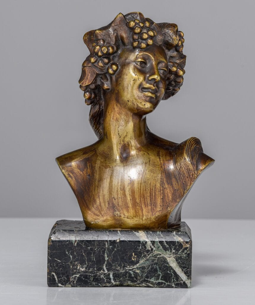 Bronzen buste van een lachende figuur, versierd met druiventrossen en bladeren op het hoofd, op een donkergroene marmeren voet tegen een effen grijze achtergrond.