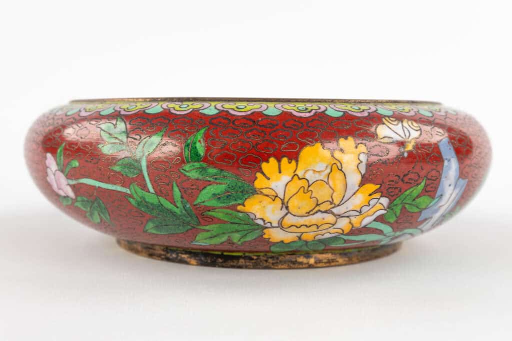 Een ronde, ondiepe kom met een rode achtergrond, versierd met kleurrijke bloemmotieven, waaronder een grote gele bloem, groene bladeren en roze en blauwe accenten. De kom heeft een licht versleten, metalen rand en voet.