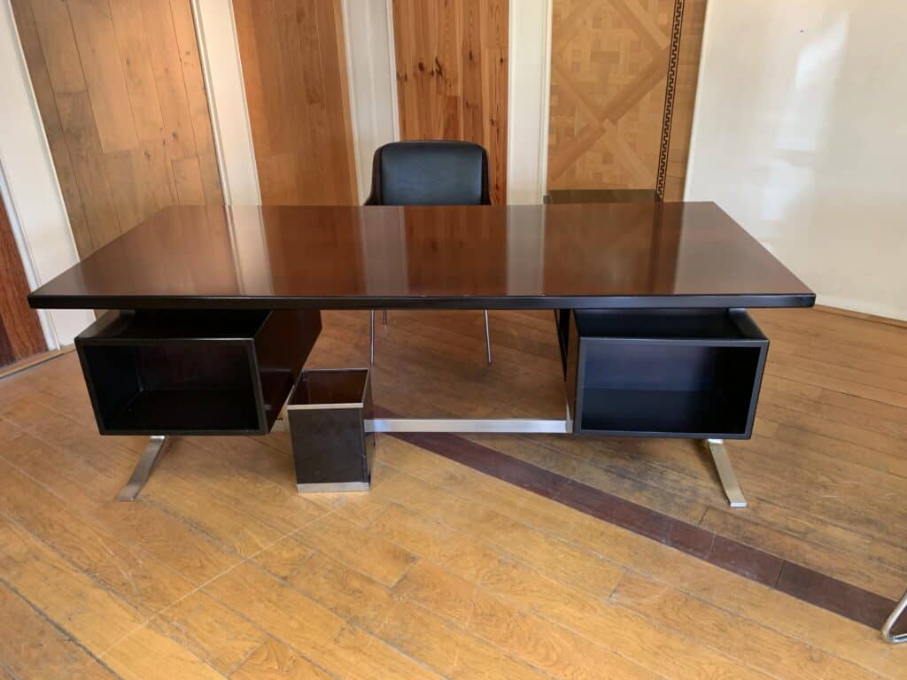 Een modern bureau van donker hout met twee open zijvakken, een zwarte prullenbak eronder en een zwarte bureaustoel erachter, op een houten vloer met lichtgekleurde houten paneelwanden.