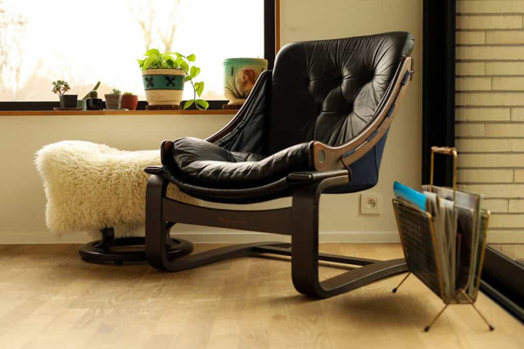 Een gezellige hoek in de woonkamer met een zwarte, gestoffeerde loungestoel, een zachte witte voetenbank, potplanten op de vensterbank en een vintage tijdschriftenrek met vinylplaten op de houten vloer.