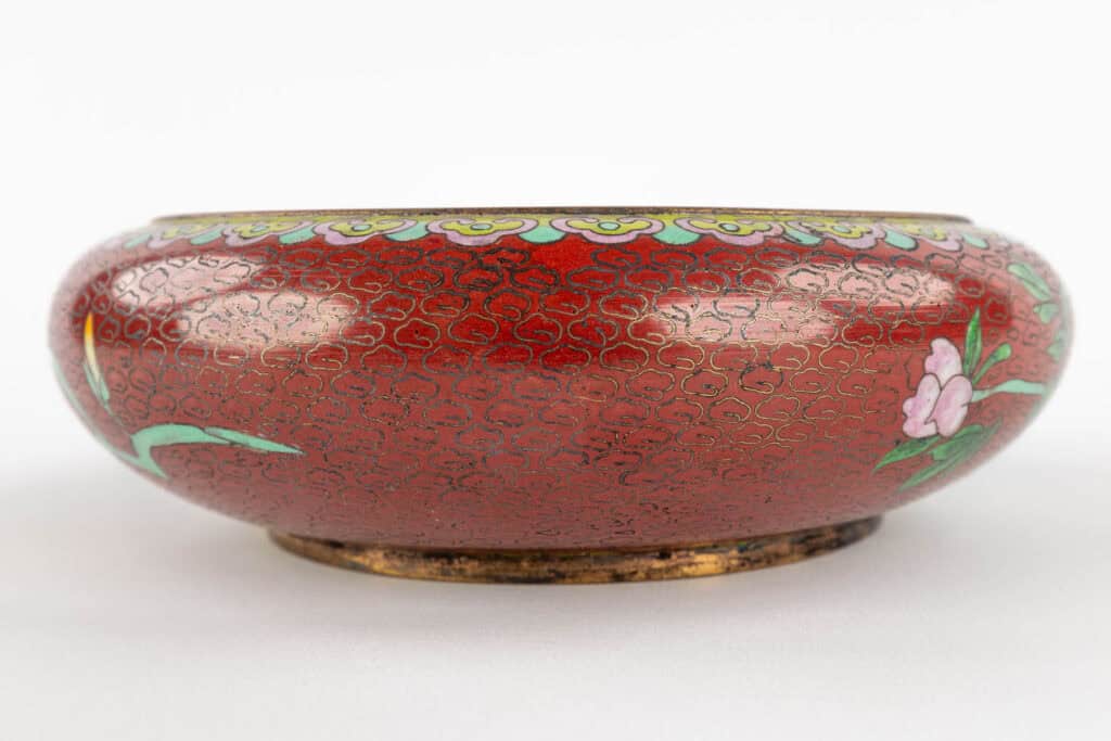 Een ronde, ondiepe, rode cloisonné kom met gouden wervelpatronen, groene bladeren en roze bloemmotieven op de zijkanten, tegen een effen witte achtergrond.