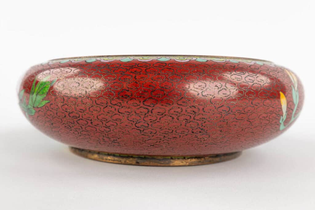 Een ronde, ondiepe rode kom met ingewikkelde zwarte patronen, een gouden rand en accenten van groene en gele decoratieve ontwerpen aan de zijkanten, rustend op een wit oppervlak.