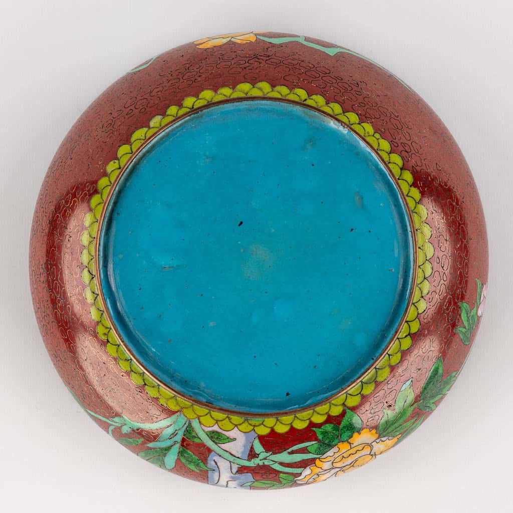 Een ronde kom van bovenaf gezien, met een helder turquoise midden, een rode rand met gele en groene gekartelde randen en bloemmotieven met groene bladeren aan de buitenrand.
