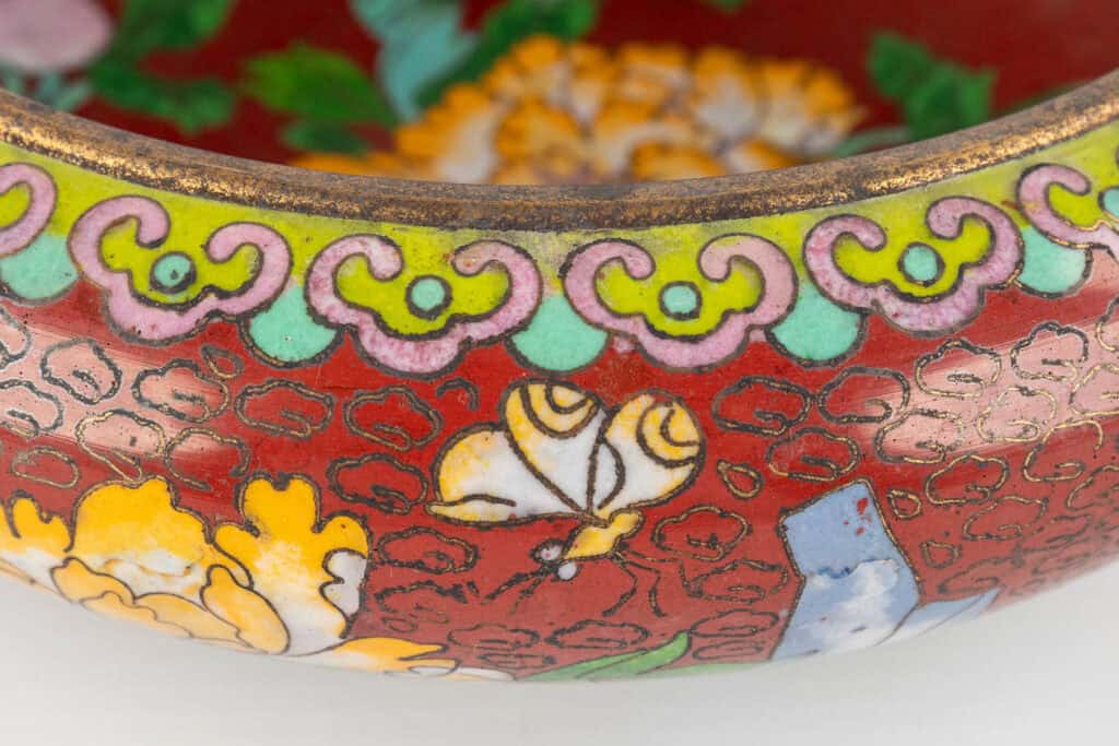 Close-up van een decoratieve kom met een rode achtergrond, met ingewikkelde bloemen- en vlinderpatronen in geel, groen, roze en wit emaille, met een herhalend gegolfd patroon aan de rand.