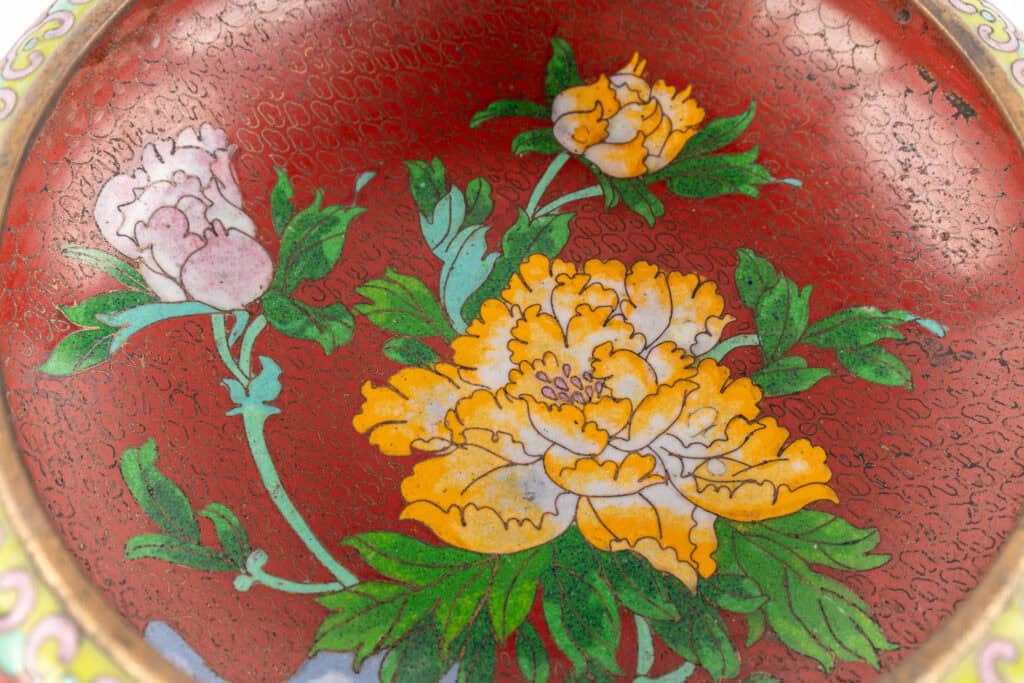 Een close-up van een decoratieve kom met een rode achtergrond, versierd met gedetailleerde gele en witte pioenrozen en groene bladeren. Het ontwerp is omlijnd met fijne zwarte lijntjes en heeft een traditionele cloisonné-emailstijl.
