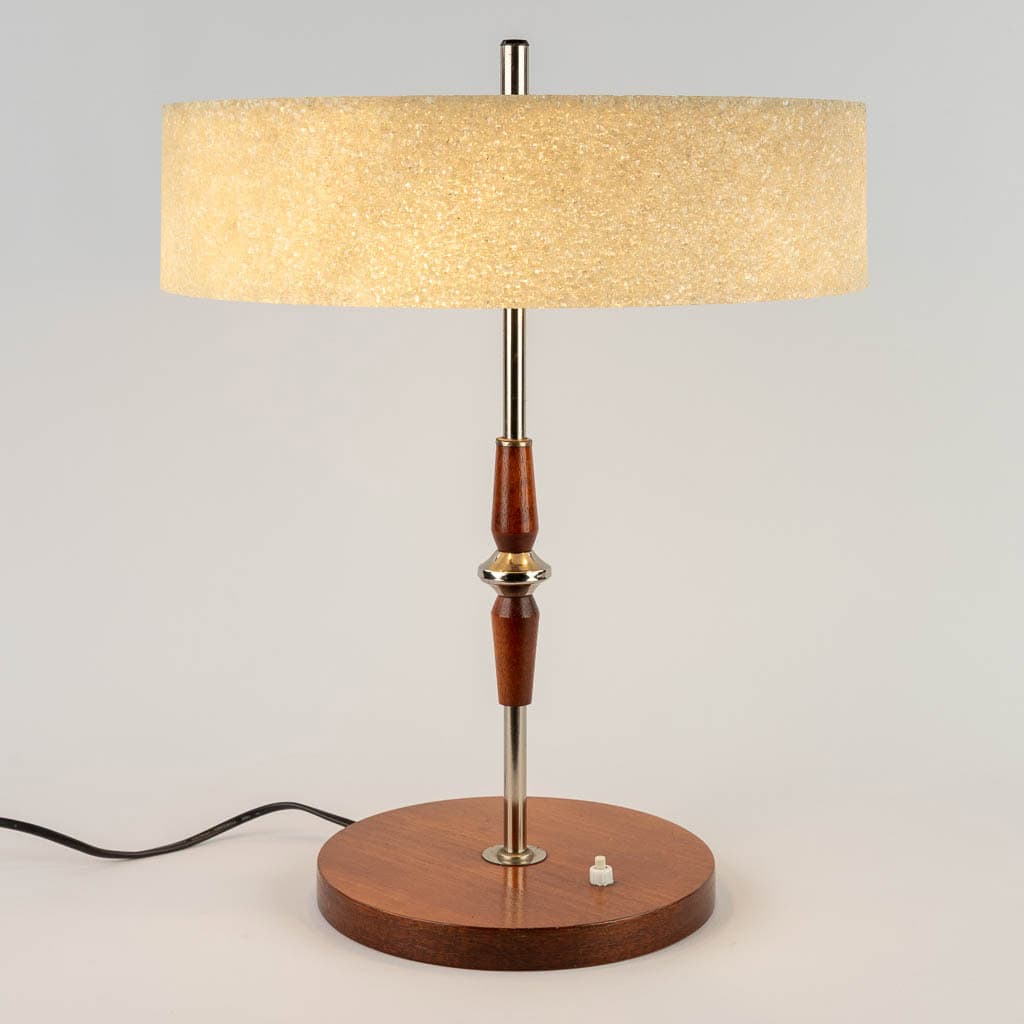 Een mid-century moderne tafellamp met een ronde houten voet, metalen accenten, een beige lampenkap met structuur en een kleine witte schakelaar op de voet. De lamp wordt aangesloten met een zwart snoer.