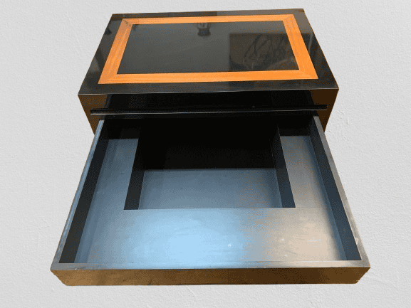 Een zwarte doos met houten afwerking en een open lade, waaruit een groot vierkant vak met een opstaande rand tevoorschijn komt. De doos staat op een lichtgekleurde ondergrond.