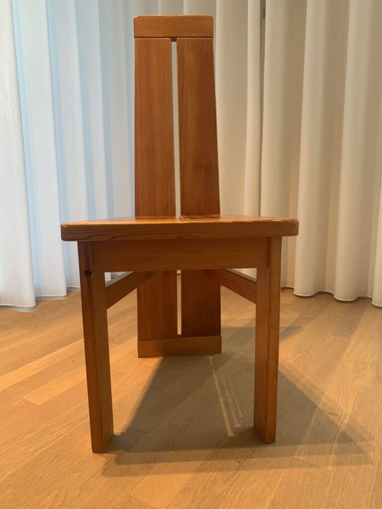 Een houten stoel met een minimalistisch, geometrisch ontwerp, voorzien van een hoge, gespleten rugleuning en schuine poten, geplaatst op een lichte houten vloer voor witte gordijnen.