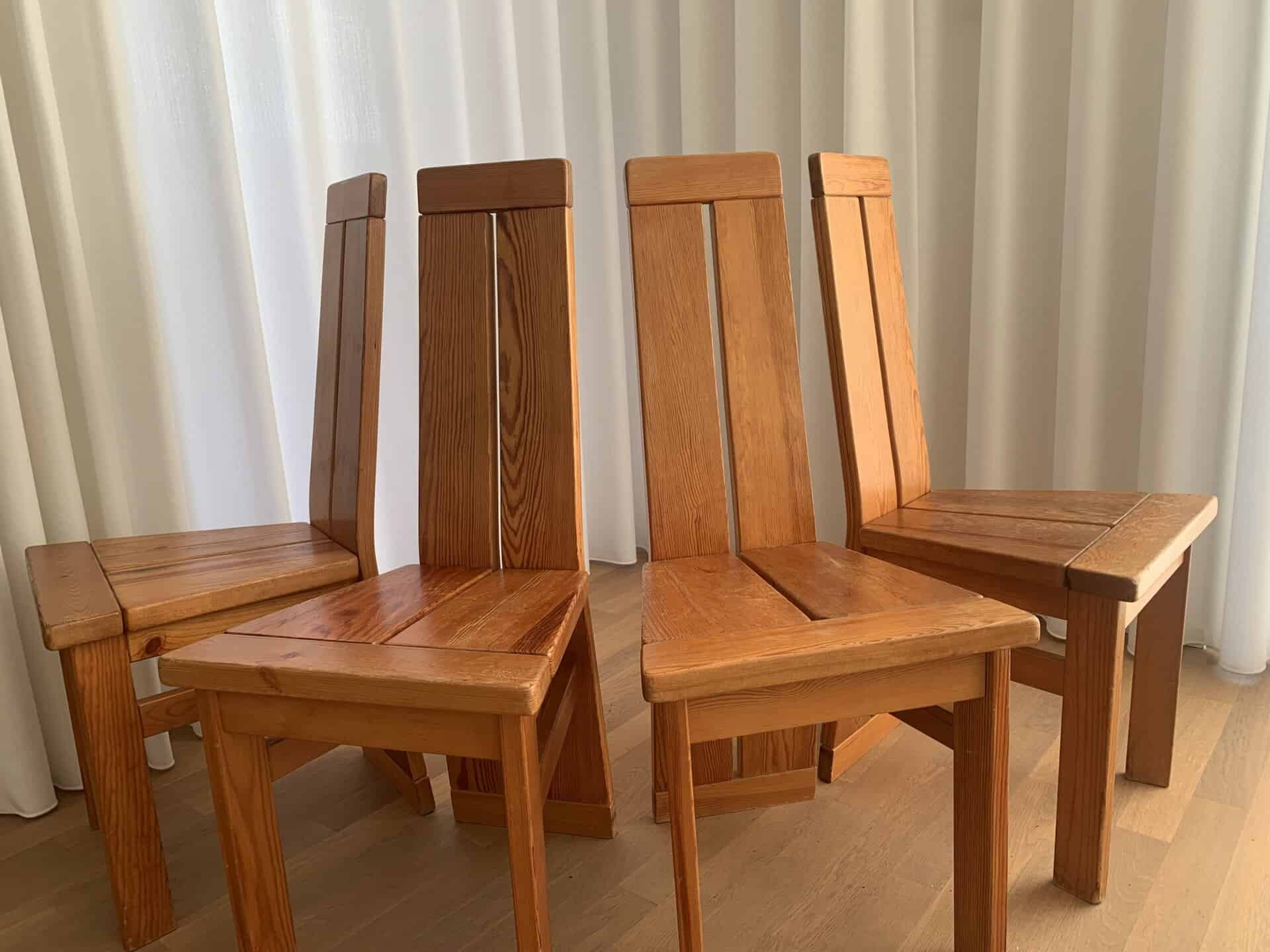 Vier houten stoelen met hoge, schuine rugleuningen staan dicht bij elkaar op een lichte houten vloer, met witte gordijnen op de achtergrond. Natuurlijk licht accentueert de nerfstructuur en warme tinten van de stoelen.