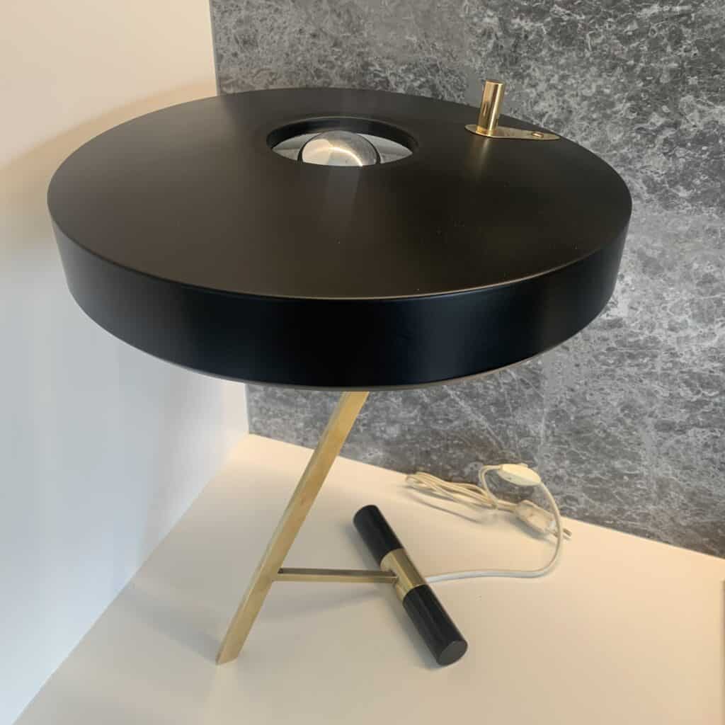Een moderne zwart-gouden tafellamp met een ronde kap, schuine gouden poten en een zichtbare gloeilamp in het midden, geplaatst op een wit oppervlak tegen een grijs-witte achtergrond.