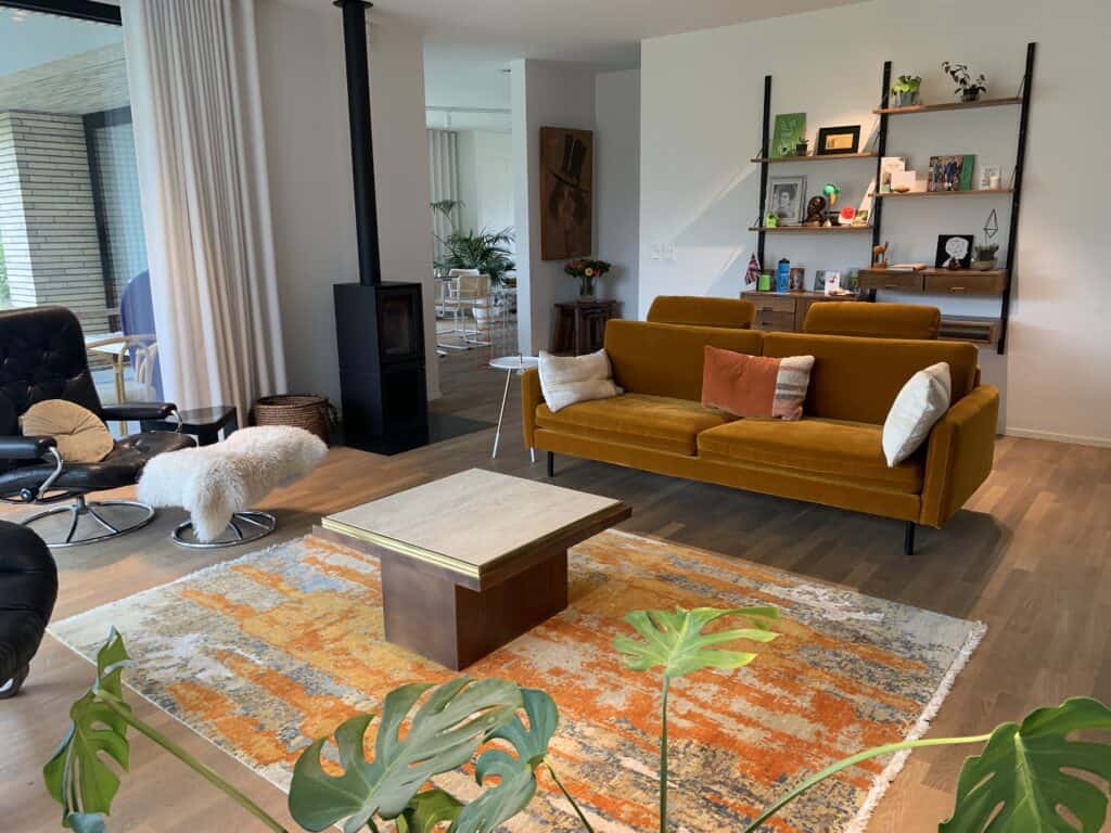 Een moderne woonkamer met een mosterdgele bank, een oranje vloerkleed, een houten salontafel, een zwarte fauteuil met een witte, zachte poef en wandplanken met boeken en decoratie. Grote ramen en planten zorgen voor natuurlijk licht.