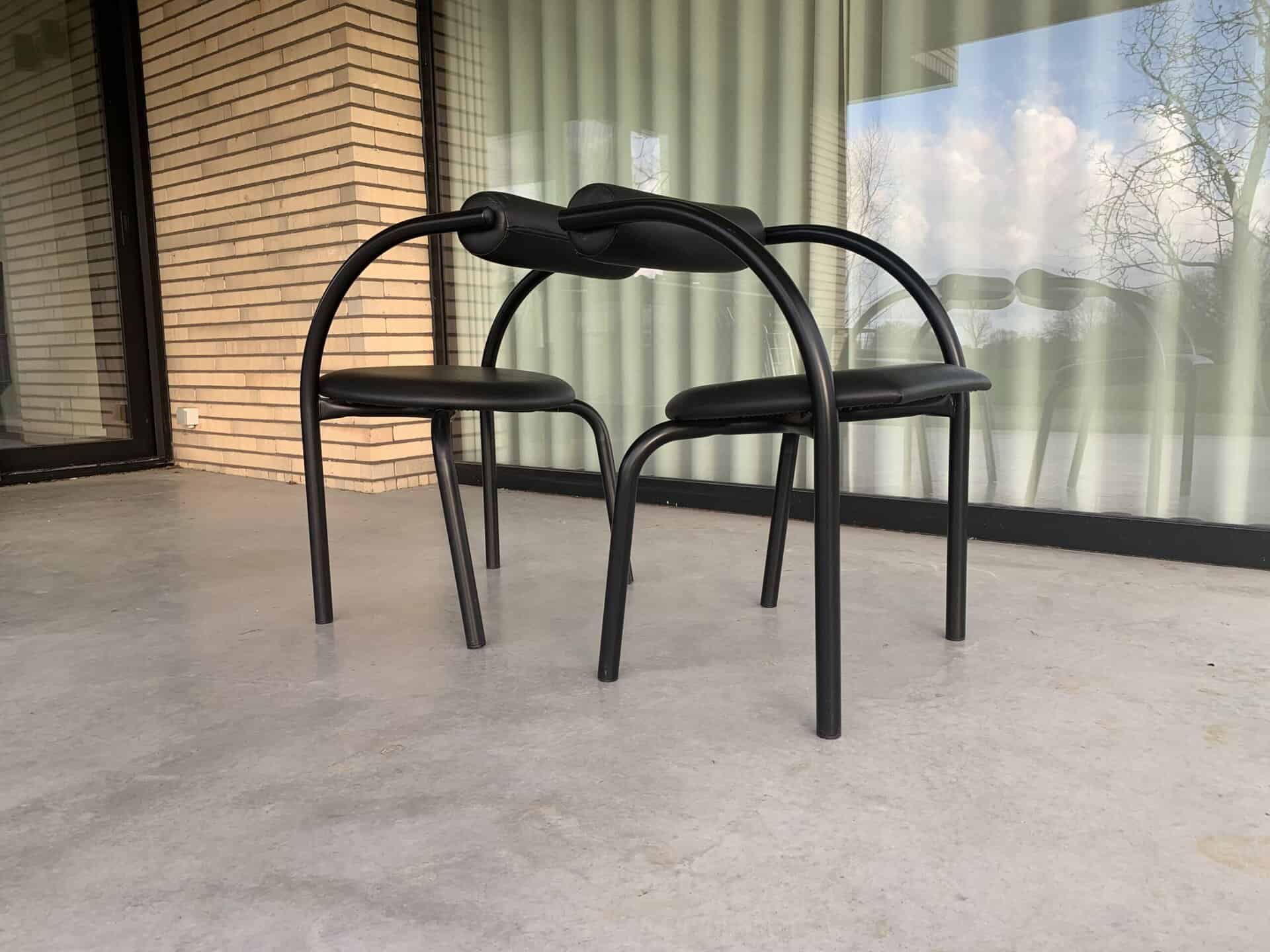 Twee moderne, zwarte stoelen met gebogen poten en rugleuningen staan rug aan rug op een betonnen terras voor een gebouw met bakstenen muren en grote glazen ramen met verticale jaloezieën.
