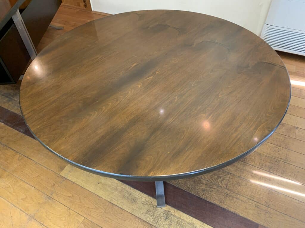 Een ronde houten tafel met een gepolijst oppervlak en zichtbare houtnerf, op een houten vloer in een goed verlichte binnenruimte.